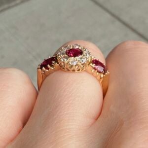 Rose gold ring with a central oval red gemstone, haloed by small diamonds and diamond-studded band on a finger