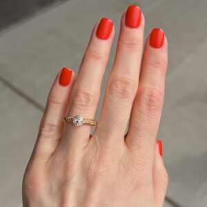 Close-up of a hand with bright orange nail polish wearing a gold ring with a diamond solitaire on the ring finger