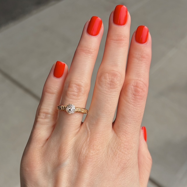 Close-up of a hand with bright orange nail polish wearing a gold ring with a diamond solitaire on the ring finger