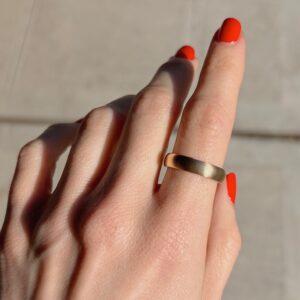 Close-up of a hand wearing a brushed gold ring on the ring finger, with bright red nail polish and sunlight casting soft shadows on the skin.