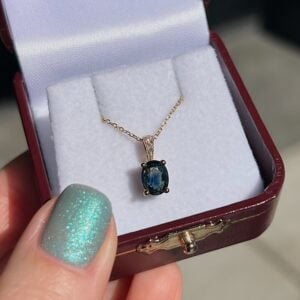 Blue oval gemstone pendant on a gold chain in an open burgundy jewelry box surface lining shows the jewelry set-up.