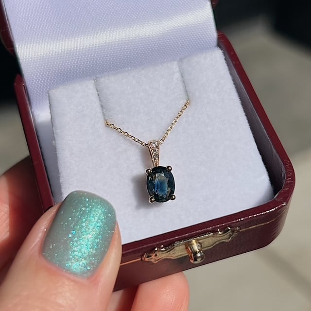 Blue oval gemstone pendant on a gold chain in an open burgundy jewelry box surface lining shows the jewelry set-up.