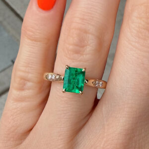 Finger adorned with a gold ring featuring a large rectangular emerald and a slim diamond-studded band on either side; orange nail polish visible.