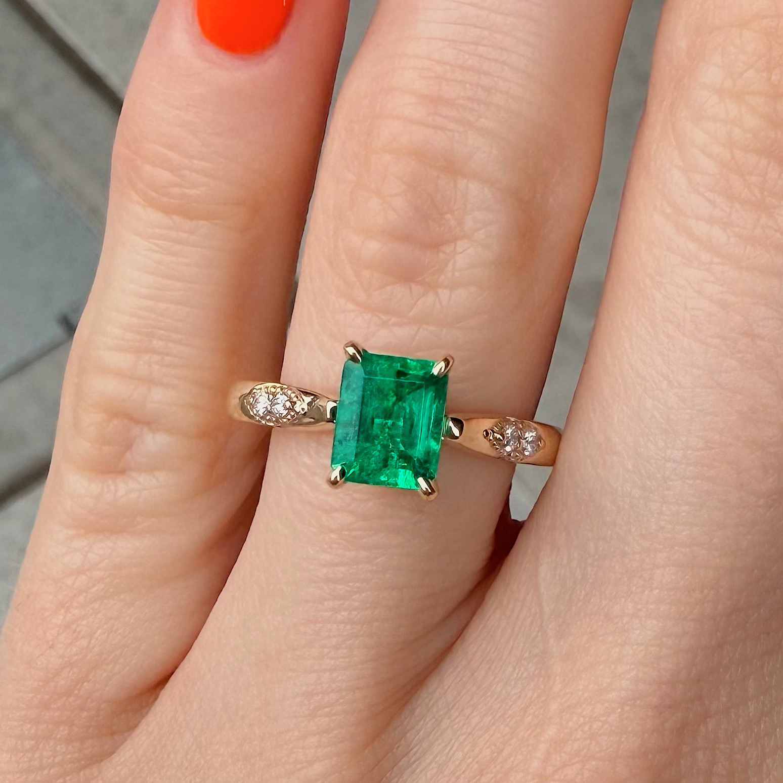 Finger adorned with a gold ring featuring a large rectangular emerald and a slim diamond-studded band on either side; orange nail polish visible.