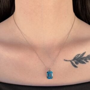 Silver chain necklace with a rectangular blue emerald-cut pendant resting on the collarbone, with a small leaf tattoo visible on the right side of the chest.