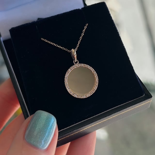 Gold circular pendant with a diamond-studded rim on a delicate chain, resting in a black jewelry box. ices