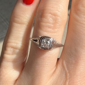 Engagement ring with a square-cut diamond on a finger with red nail polish in sunlight, showing a silver band and bezel setting