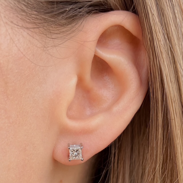 Close-up of an ear with a small square-cut diamond stud earring in a silver setting on the earlobe.