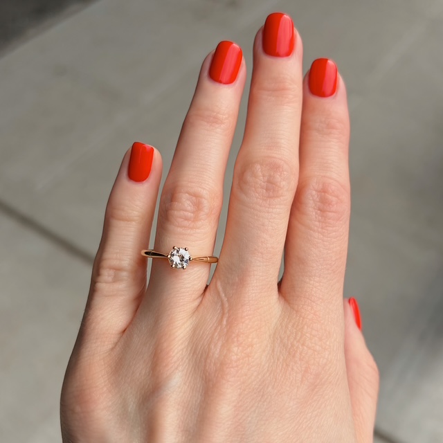 Close-up of a hand with bright red nails wearing a gold solitaire ring featuring a single clear gemstone on the ring finger (left hand).
