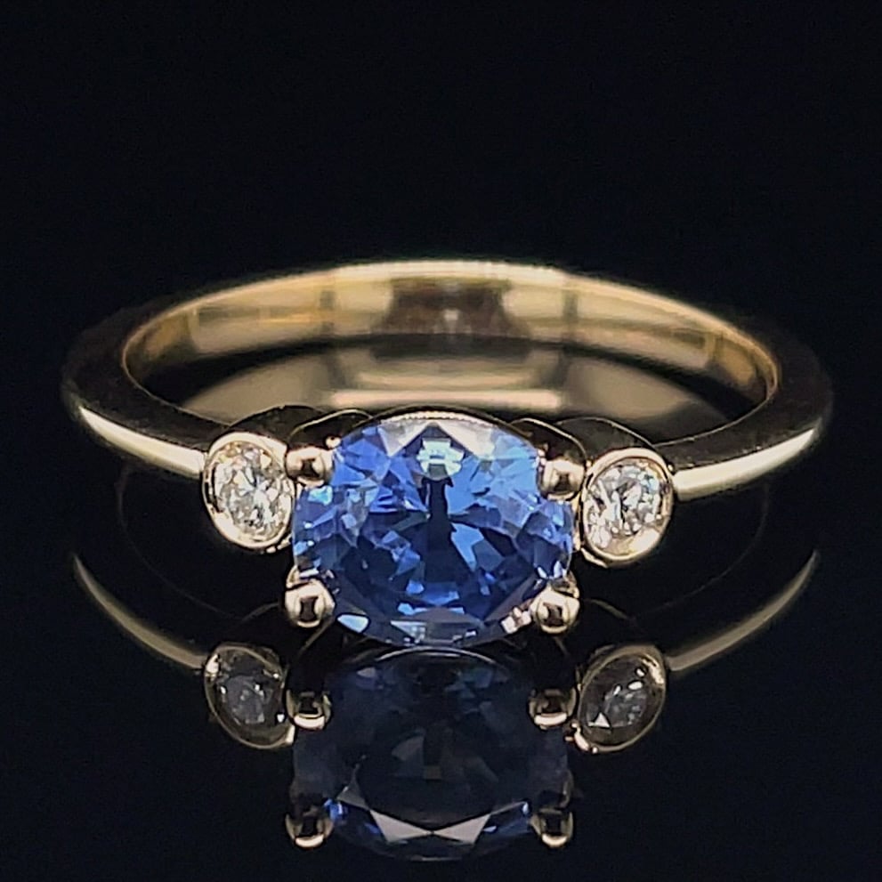 Gold ring with a large blue oval central gemstone and two diamonds on the sides, set on a reflective black surface.
