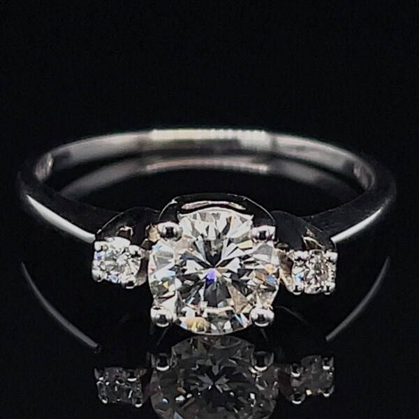 Engagement ring with a large central diamond and two smaller side diamonds on a silver band against a black background, reflected below. Informs about a diamond ring setting and style.