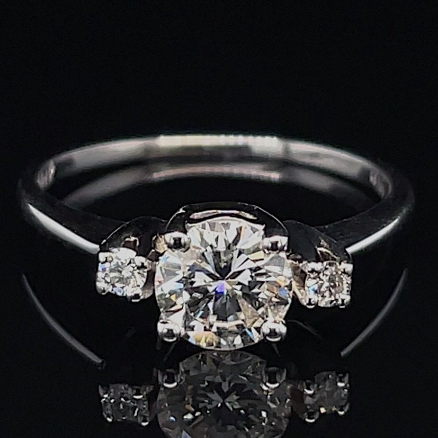 Engagement ring with a large central diamond and two smaller side diamonds on a silver band against a black background, reflected below. Informs about a diamond ring setting and style.