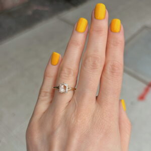 Hand with bright yellow manicured nails wearing a gold ring with a hexagonal pale gemstone on the ring finger, against a sidewalk background.