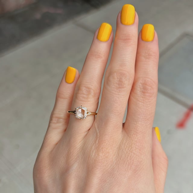 Hand with bright yellow manicured nails wearing a gold ring with a hexagonal pale gemstone on the ring finger, against a sidewalk background.