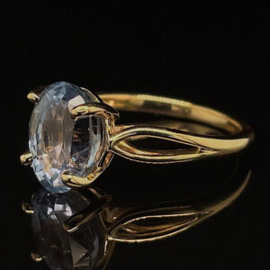 Gold ring with an oval-cut light blue gemstone held by four prongs against a black background, reflection visible beneath.