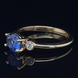 Gold ring with a large blue sapphire centerpiece and two small diamonds on the band, reflecting on a black surface