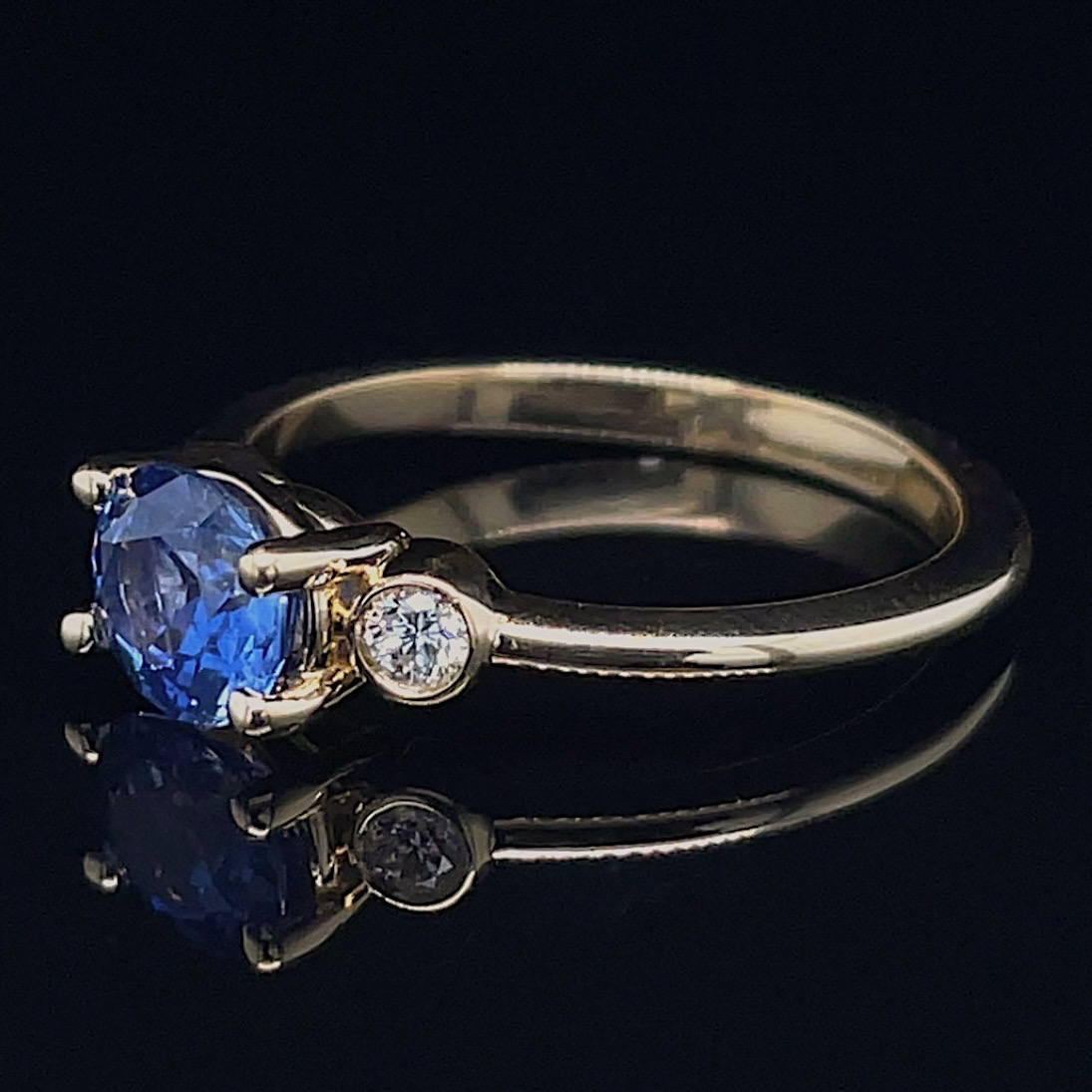 Gold ring with a large blue sapphire centerpiece and two small diamonds on the band, reflecting on a black surface
