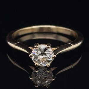 Gold solitaire engagement ring with a round diamond on a black surface, its reflection beneath the gem.