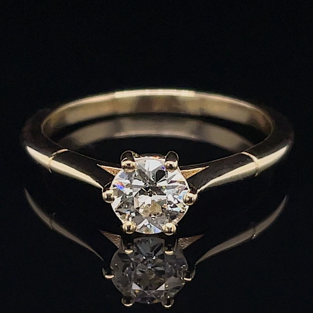 Gold solitaire engagement ring with a round diamond on a black surface, its reflection beneath the gem.