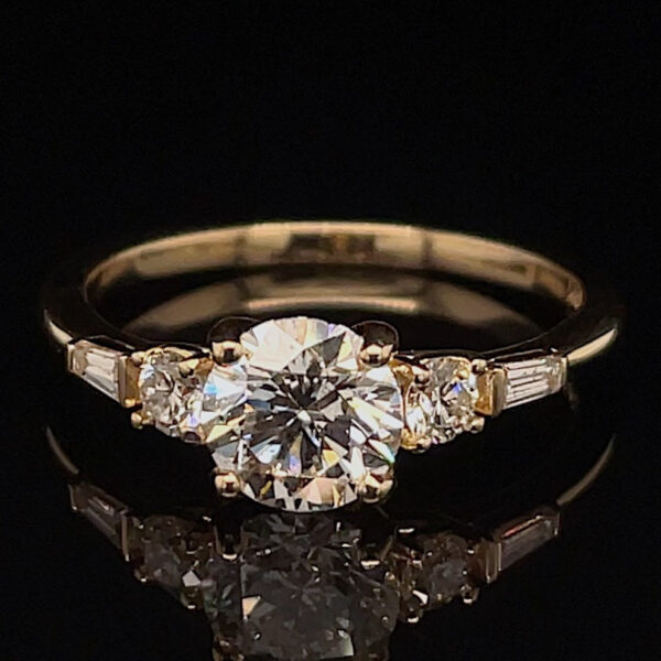 Engagement ring with a large central round-cut diamond flanked by two smaller diamonds on a gold band, resting on a black reflective surface.