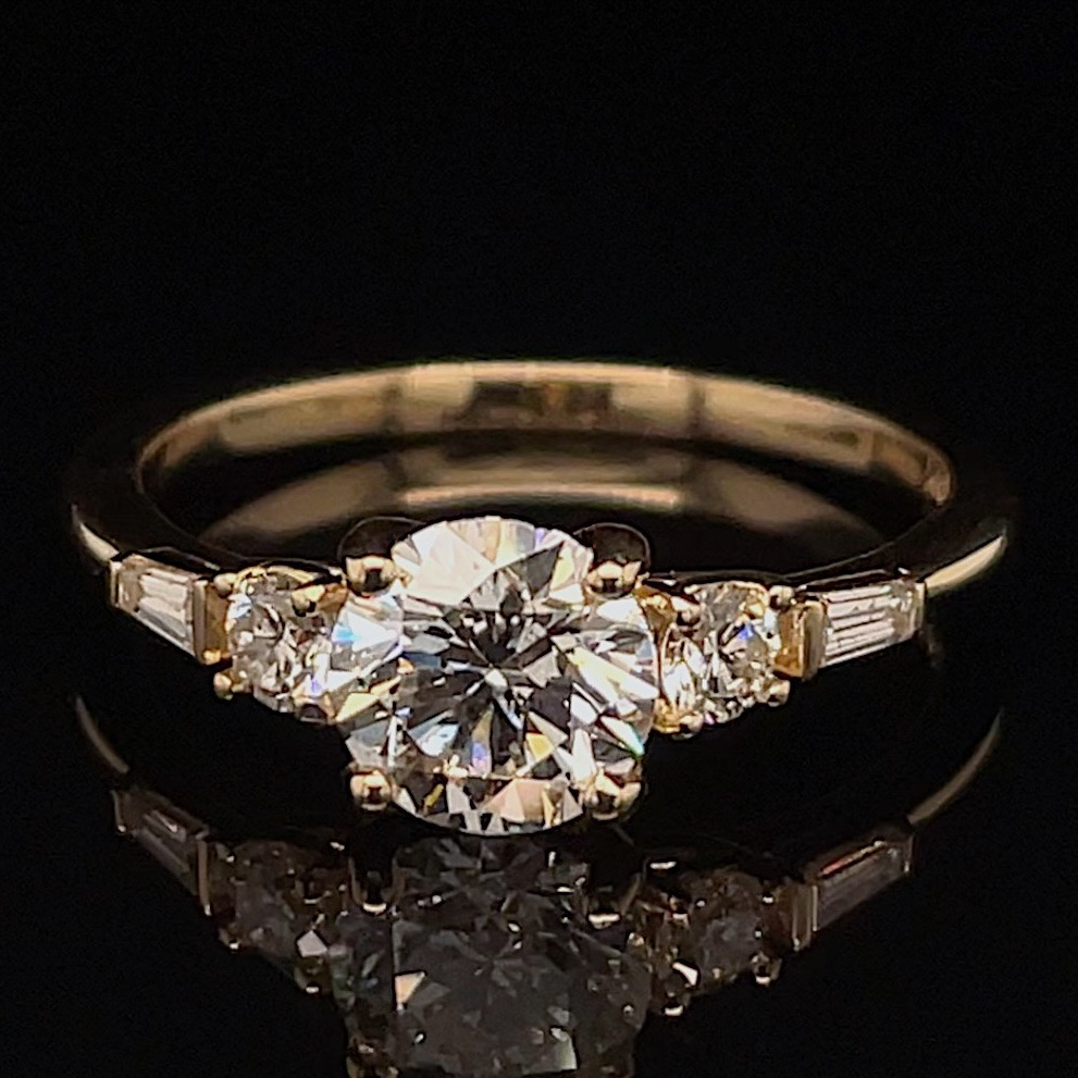Engagement ring with a large central round-cut diamond flanked by two smaller diamonds on a gold band, resting on a black reflective surface.