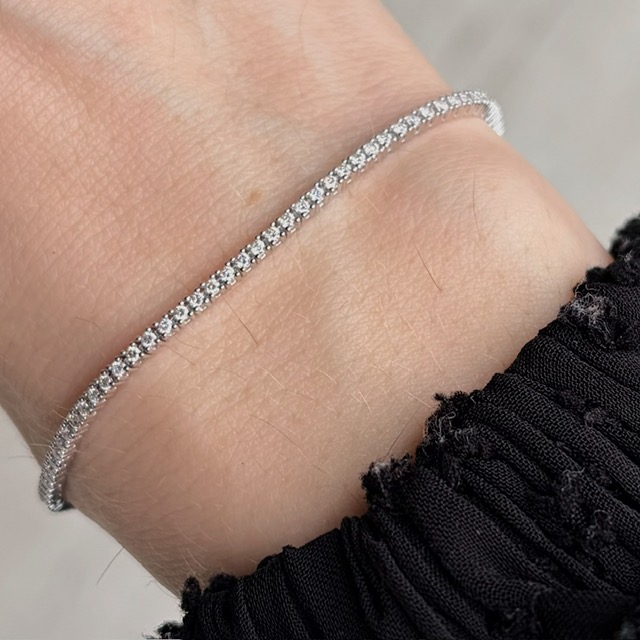 Slim diamond tennis bracelet resting on a light-skinned wrist, with a black fabric cuff nearby.
