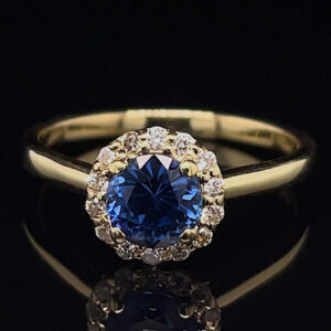 Gold ring with a deep blue oval-cut sapphire surrounded by a halo of diamonds on a dark background.