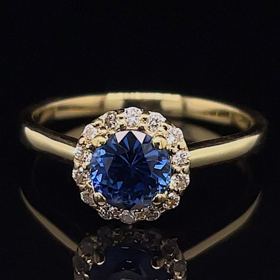 Gold ring with a deep blue oval-cut sapphire surrounded by a halo of diamonds on a dark background.