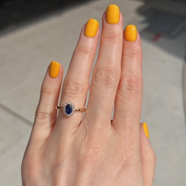 Hand with bright yellow nail polish wearing a gold ring with a blue oval gemstone encircled by small diamonds.