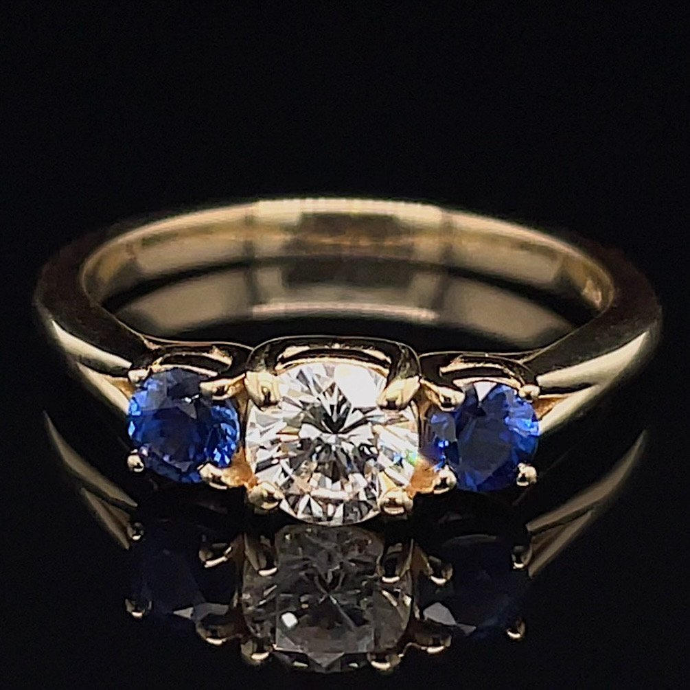 Gold ring with a central round diamond flanked by two blue sapphires on a dark background, showing reflection beneath.