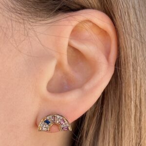 Crescent-shaped gold earring with multicolored gems on a person’s earlobe, viewed close-up from the side of the head, rainbow-styled jewelry.