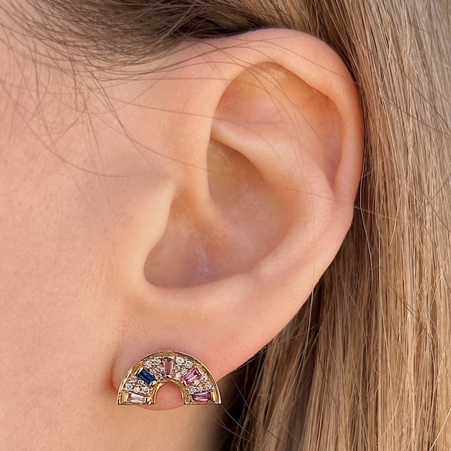 Crescent-shaped gold earring with multicolored gems on a person’s earlobe, viewed close-up from the side of the head, rainbow-styled jewelry.
