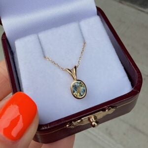 Gold pendant necklace with a pale blue gemstone in a small jewelry box, chain draped over a white cushion.