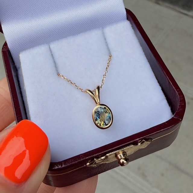 Gold pendant necklace with a pale blue gemstone in a small jewelry box, chain draped over a white cushion.