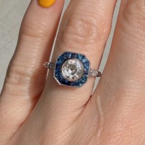 Diamond ring with blue halo stones surrounding a central diamond, worn on a finger.