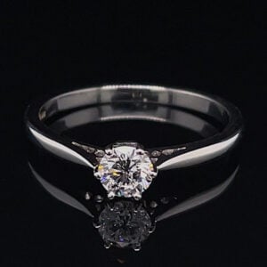 Engagement ring with a round-cut center diamond on a silver band against a black background, reflecting below it.