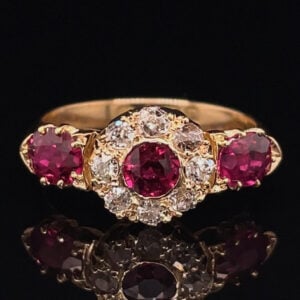 Gold ring with a central oval red ruby surrounded by diamonds, flanked by two red rubies, on a black mirrored surface.