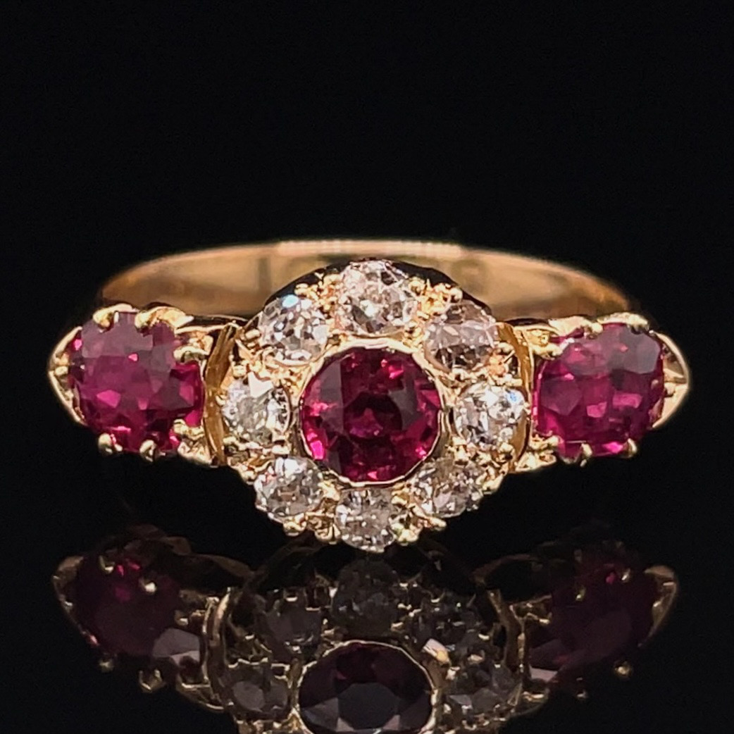 Gold ring with a central oval red ruby surrounded by diamonds, flanked by two red rubies, on a black mirrored surface.