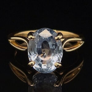 Gold ring with a large oval clear gemstone set in a four-prong gold band, against a black reflective surface.
