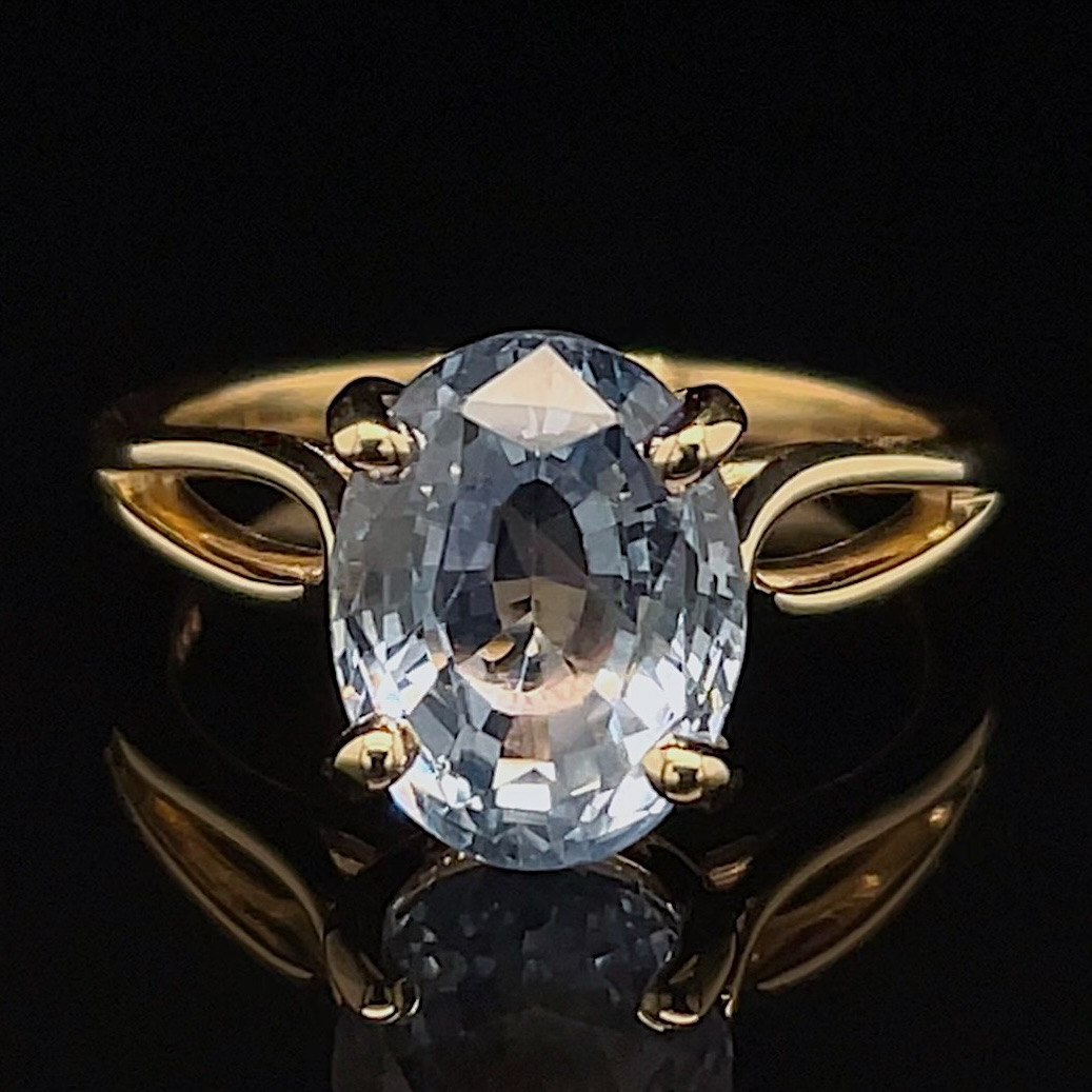 Gold ring with a large oval clear gemstone set in a four-prong gold band, against a black reflective surface.