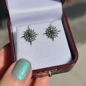 Starburst stud earrings with green center stones and surrounding diamonds in a white cushioned jewelry box.