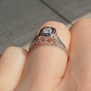 Close-up of a silver ring with a square diamond on a finger, showing detailed engraved band craftsmanship in a soft-focus background.