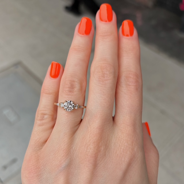 Hand with bright orange nail polish wearing a diamond engagement ring on the ring finger (close-up).