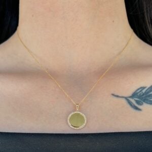 Gold chain necklace with a round gold pendant bordered by small diamonds resting on the collarbone; a blue feather tattoo is visible on the right.