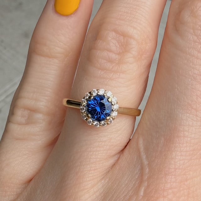 Gold ring with a deep blue center gemstone surrounded by small diamonds on a slender band, worn on a finger.