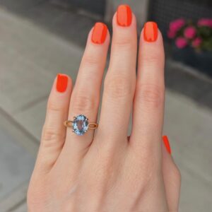 Close-up of a hand with bright orange nail polish wearing a gold ring with a blue oval gemstone on the ring finger.