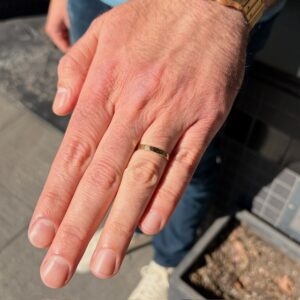 Close-up of a man’s left hand wearing a gold wedding band on the ring finger, outdoors.