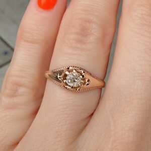 Rose gold ring with a flower-shaped setting and a central diamond on a finger with orange nail polish.