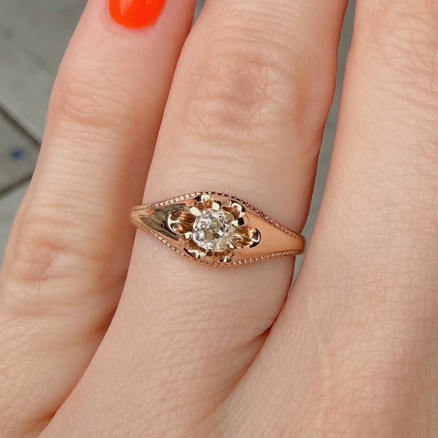 Rose gold ring with a flower-shaped setting and a central diamond on a finger with orange nail polish.
