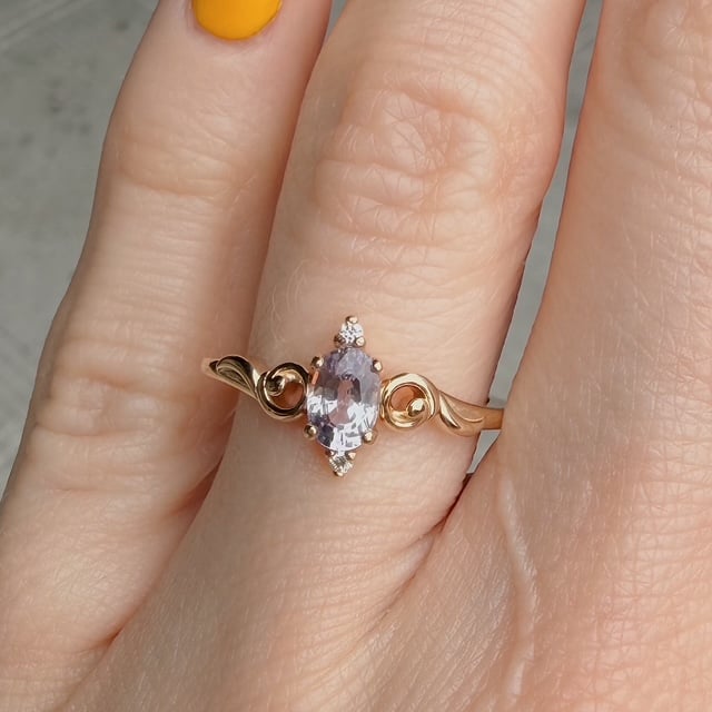 Gold ring with an oval lavender gemstone on a finger, featuring decorative gold swirls on the band.